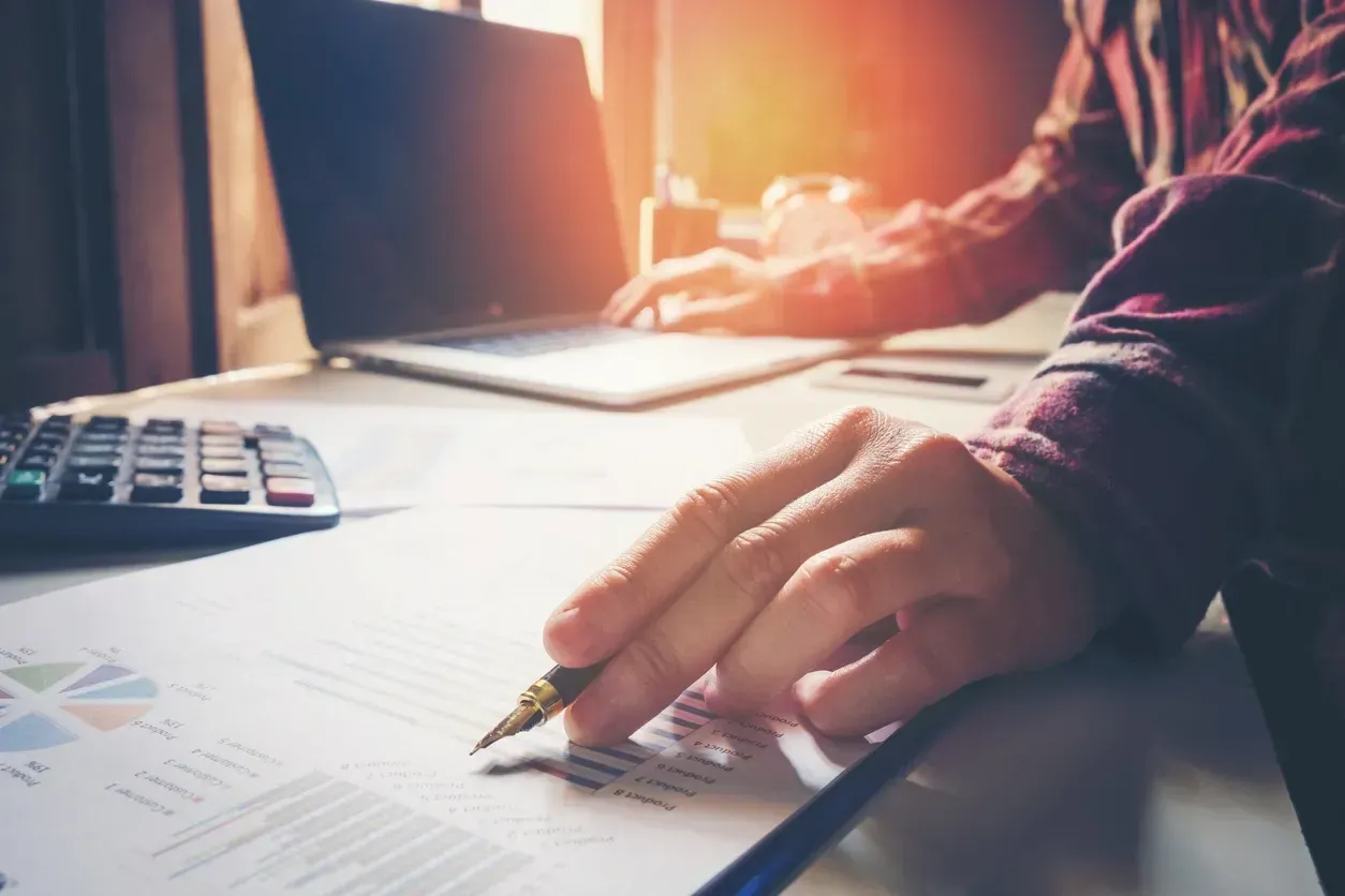 Person working on a laptop and reviewing paperwork with a pen. Focus on hands and documents.
