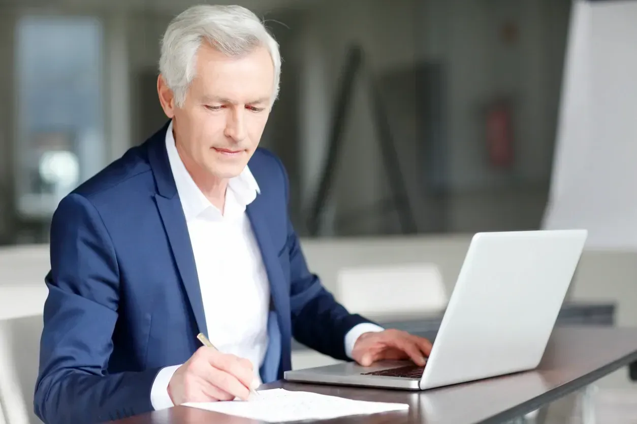 A man in a blue suit works on a laptop and writes on paper at a desk indoors.