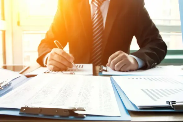 Man in a suit writing at a desk, reviewing documents in an office setting with a bright light.