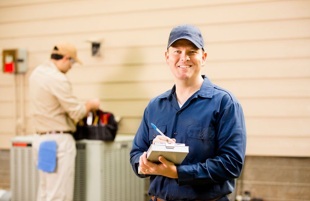 An HVAC technician in a blue shirt is holding a clipboard and smiling.