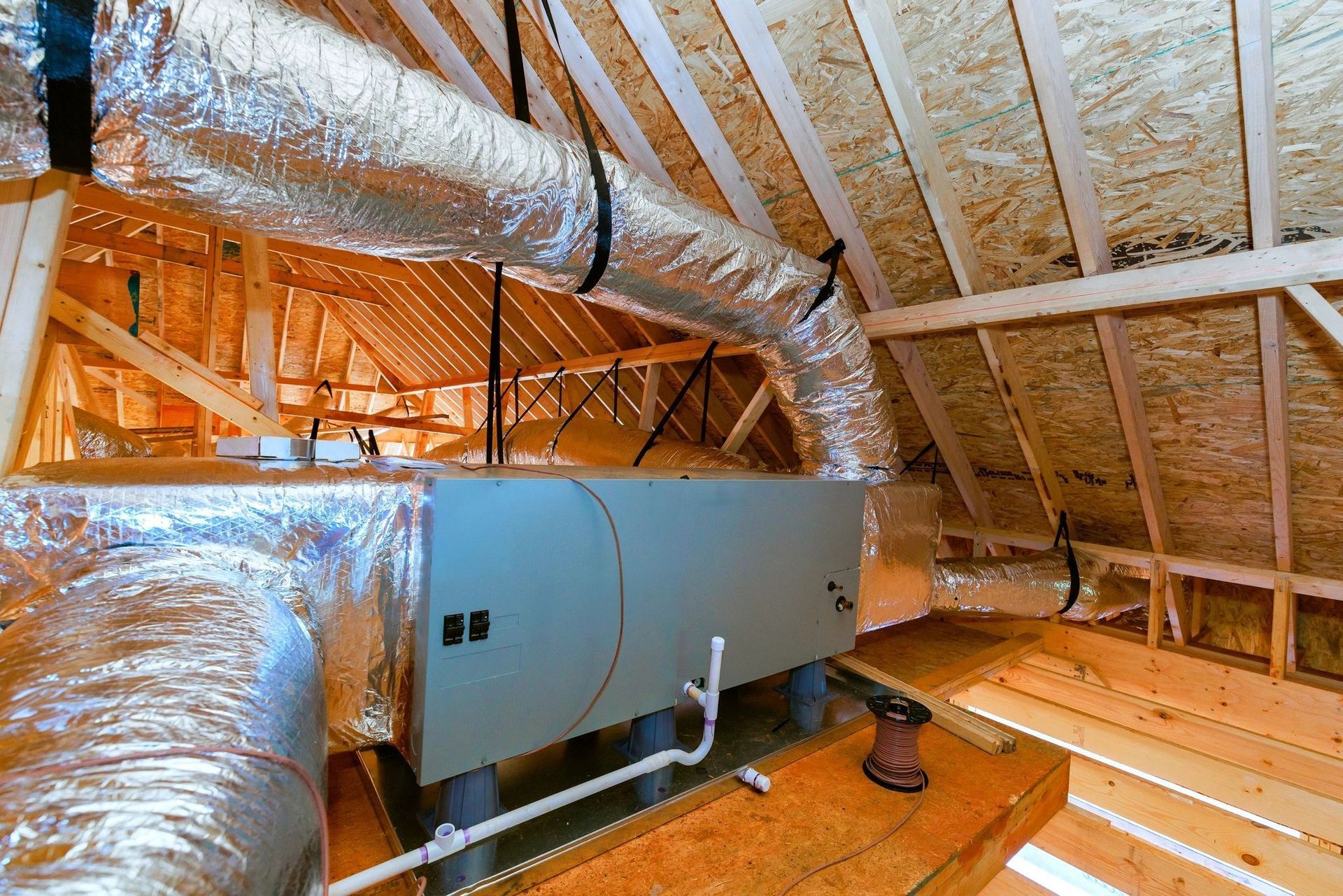 Foil-wrapped HVAC ducts and a furnace in an unfinished attic with exposed wooden rafters.