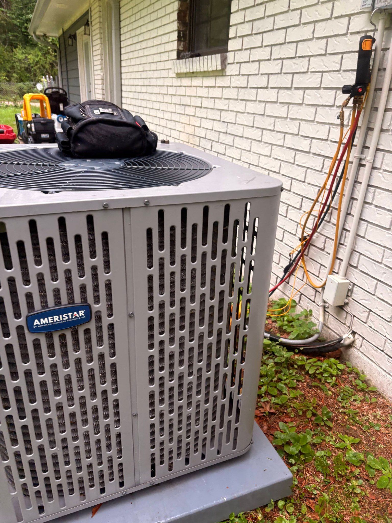 Outdoor air conditioner unit beside white brick wall, with an orange ladder and black bag nearby