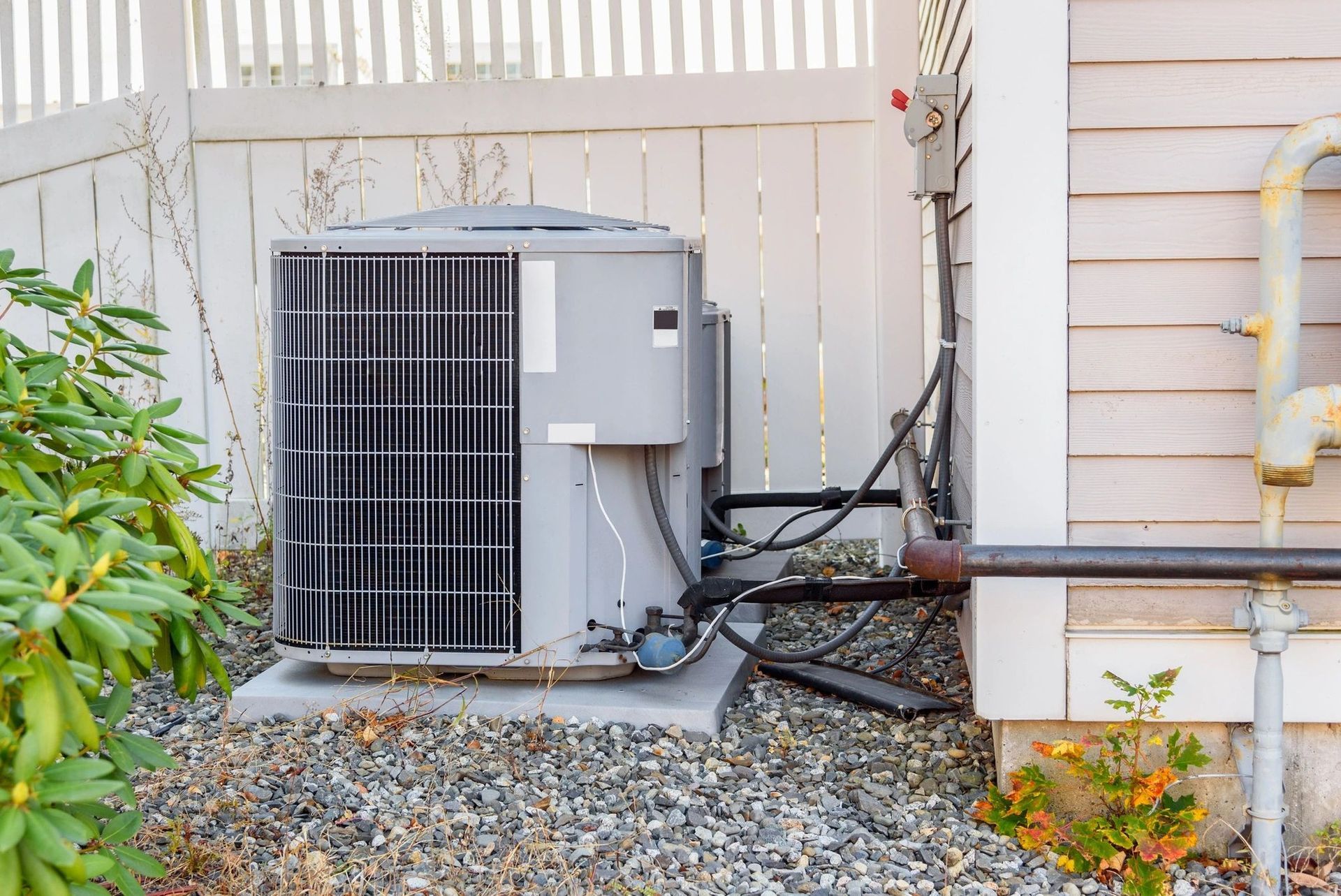 Outdoor air conditioning unit beside a house, with hoses and gravel in a fenced yard