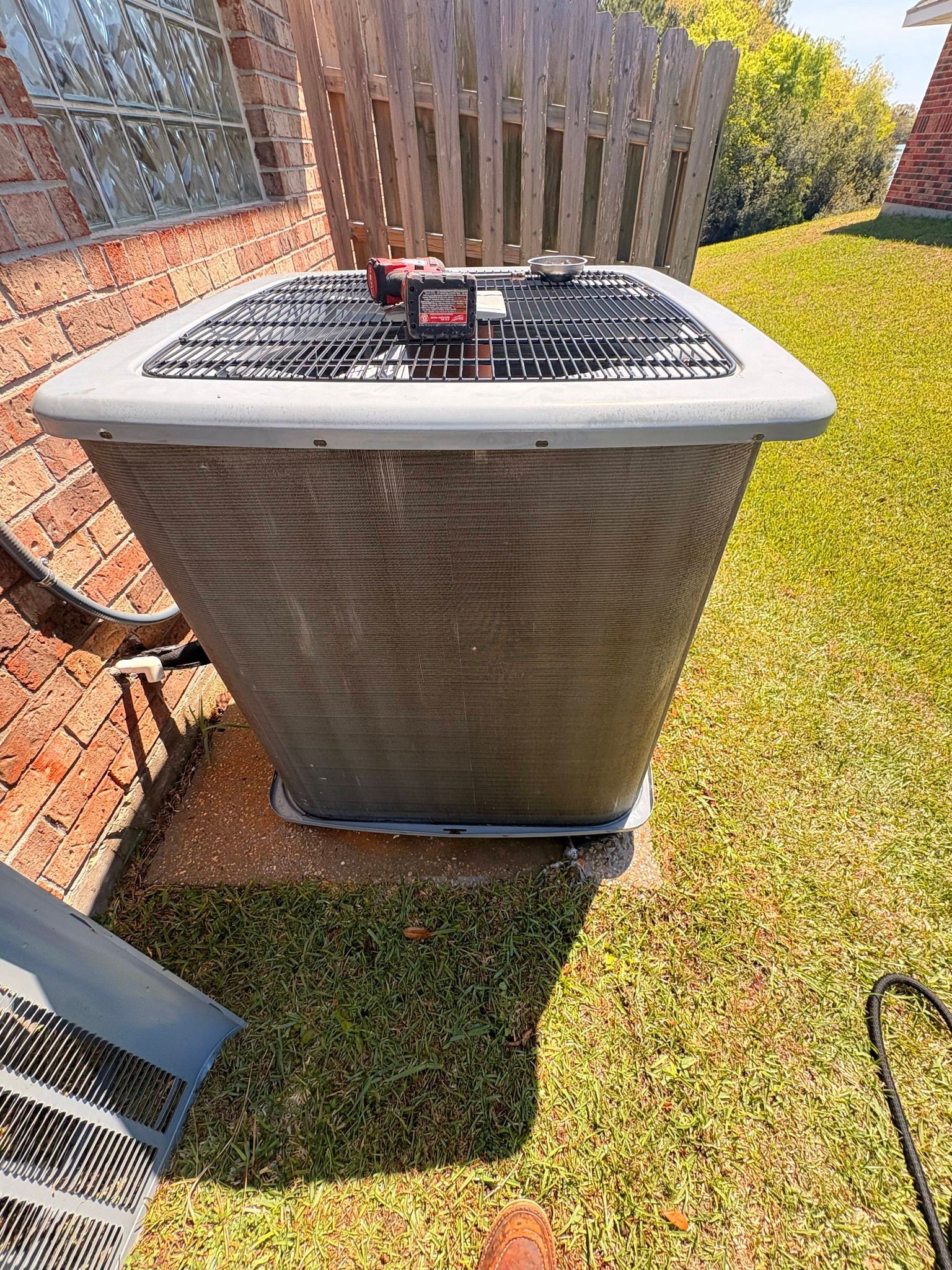 Outdoor air conditioner unit beside a wooden fence on a grassy lawn, with a small red device on top.