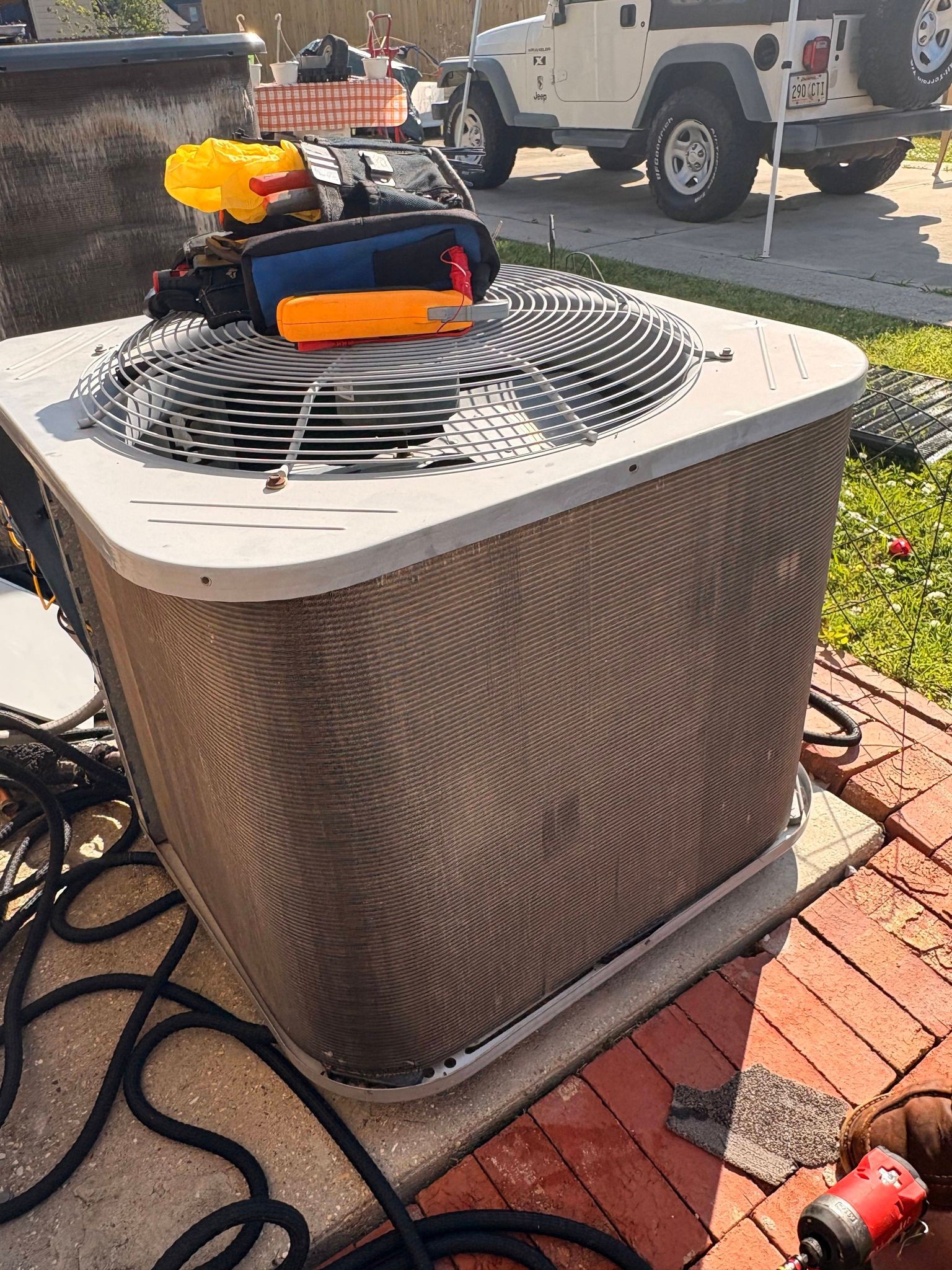 Outdoor HVAC condenser with tools and a hose on top, sitting on a brick patio near a truck.
