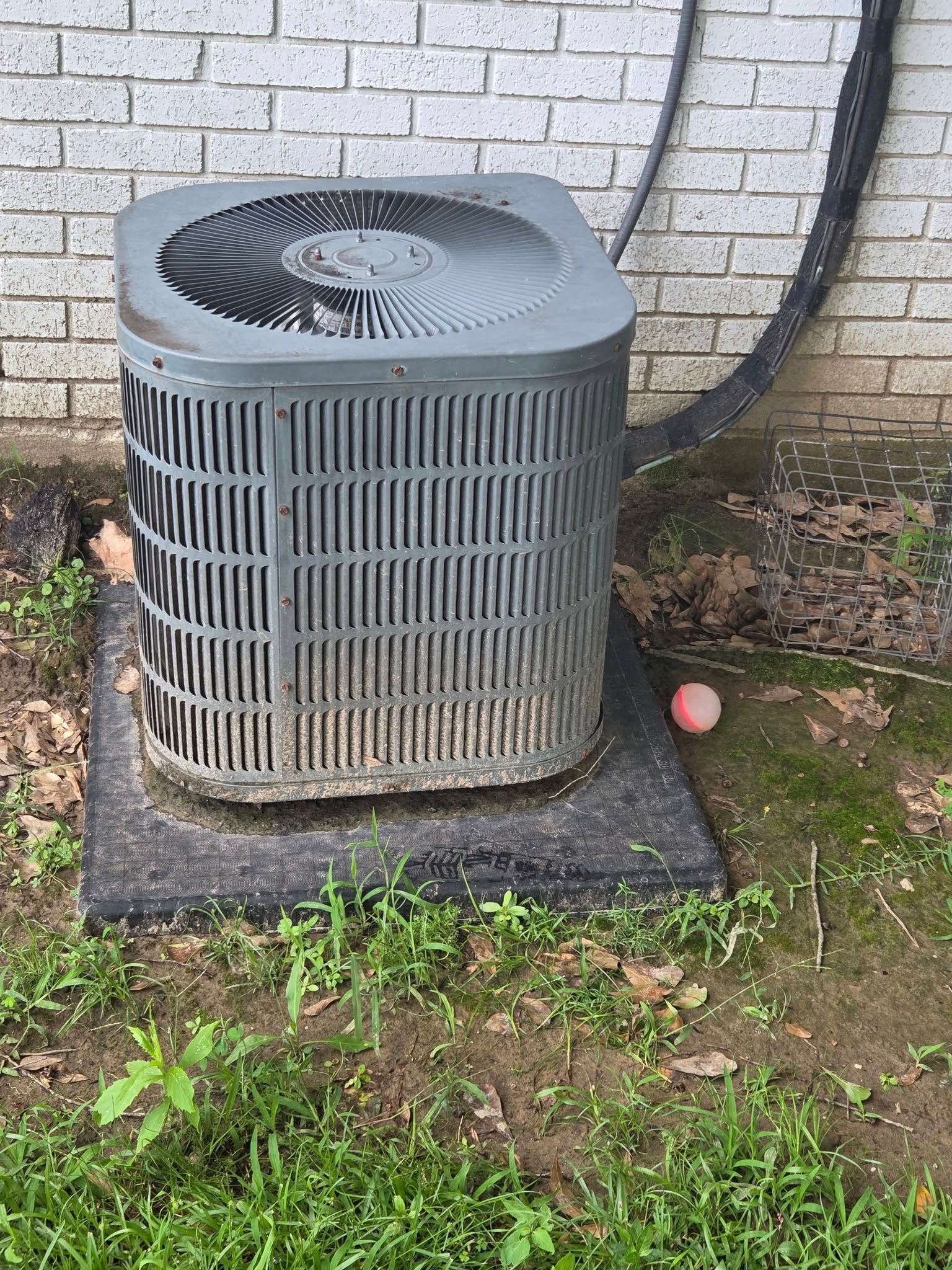 Outdoor air conditioner unit on a concrete pad beside a brick wall, with grass and a small pink ball nearby.