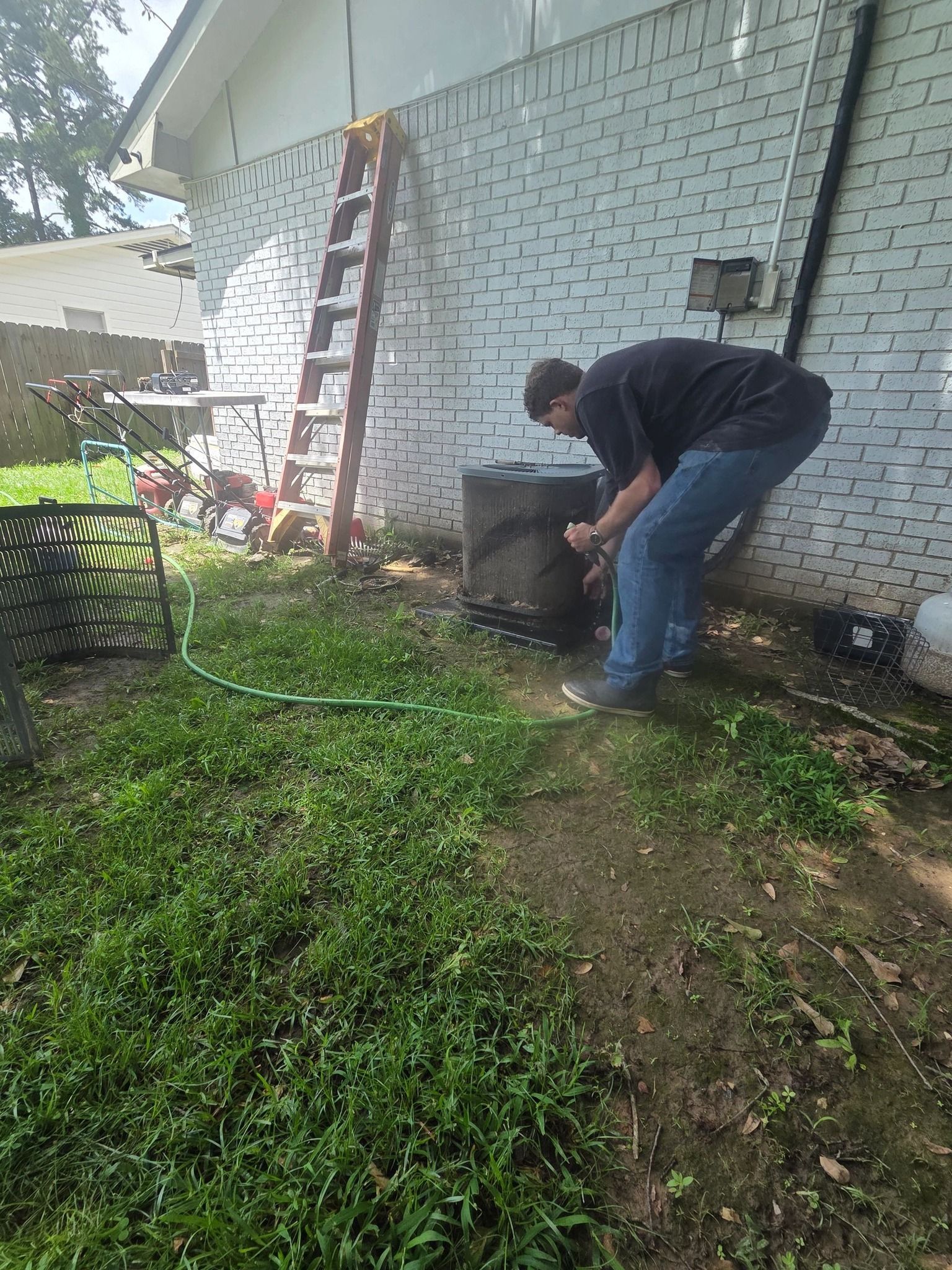 Person bending over near a brick wall, working beside a ladder and tire in a grassy yard.