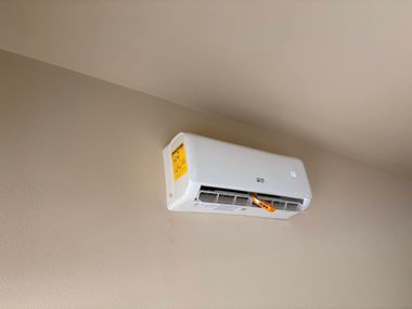 White wall-mounted air conditioner on a beige wall, with vents open and a yellow label on the side