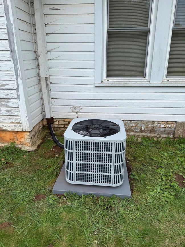 Outdoor air conditioner unit beside a white house wall and window on a small concrete pad.