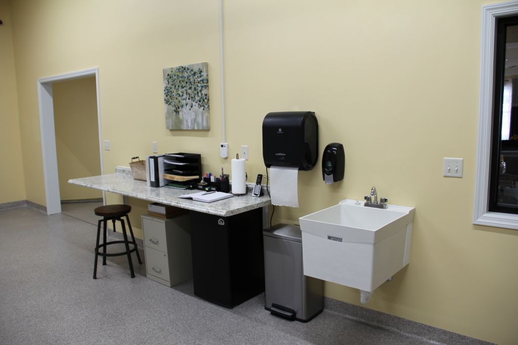 A room with a desk , sink , trash can and stool.