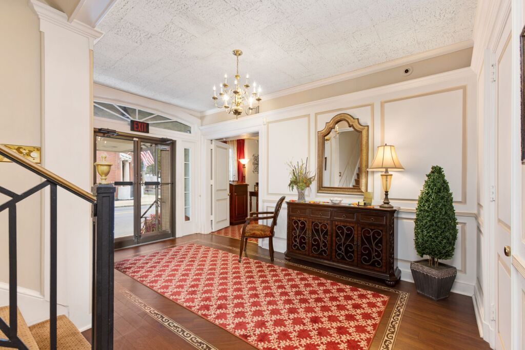 A large hallway with a red rug , stairs , a mirror and a chandelier.