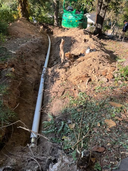 A trench in the ground with a white pipe. A green excavator and trees are in the background.