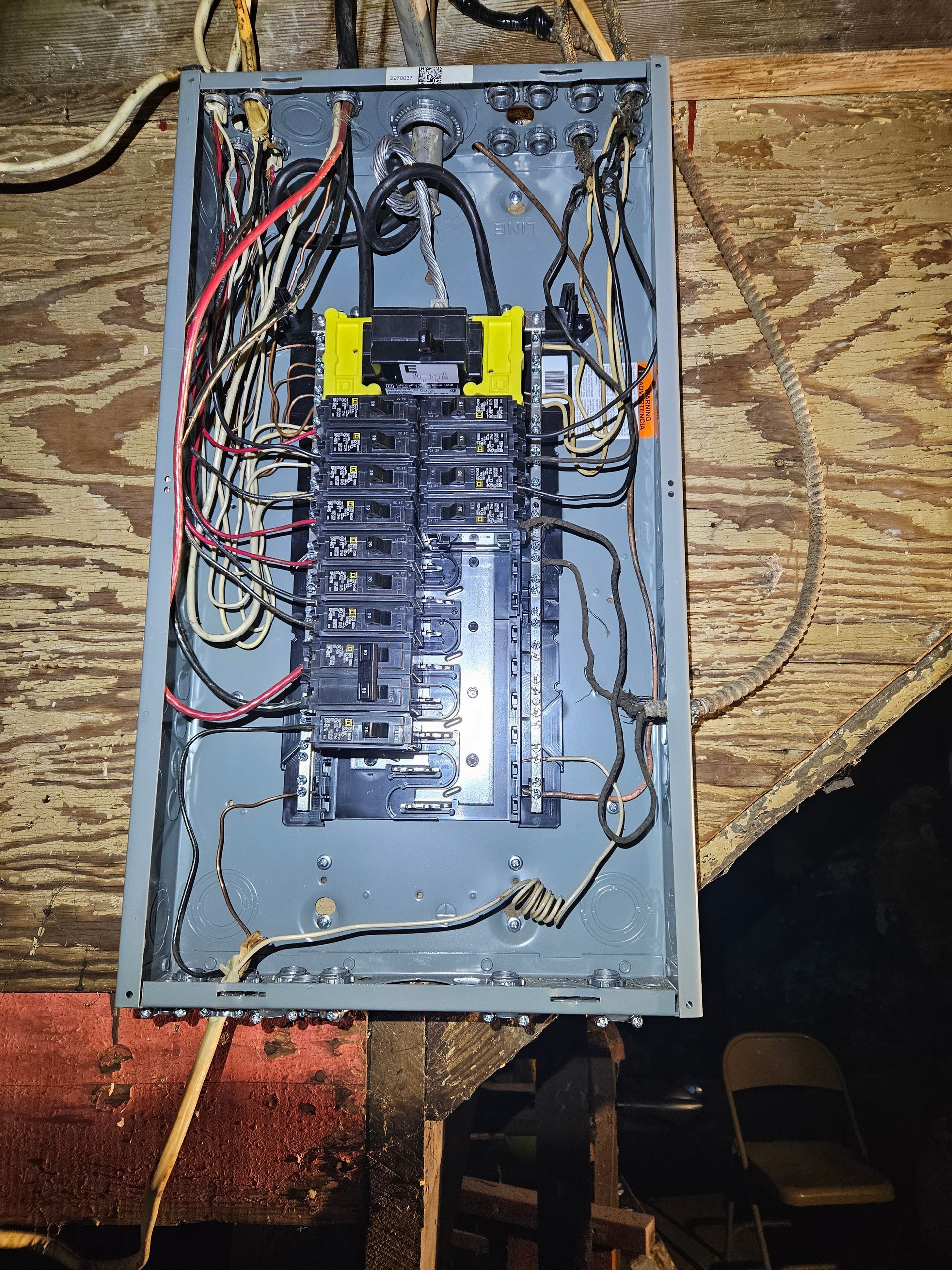 Open electrical panel with numerous wires and circuit breakers. Gray metal box mounted on wood.
