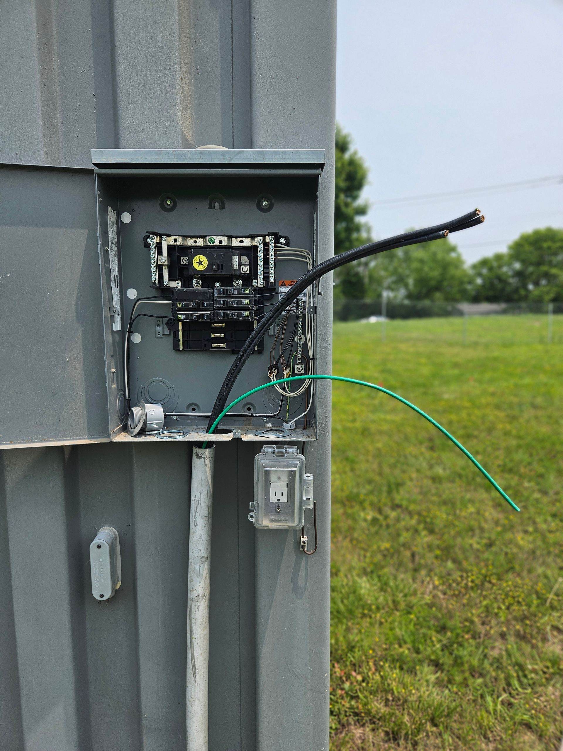 Electrical panel with exposed wires, mounted on a gray metal structure outdoors. Green and black wires are visible.
