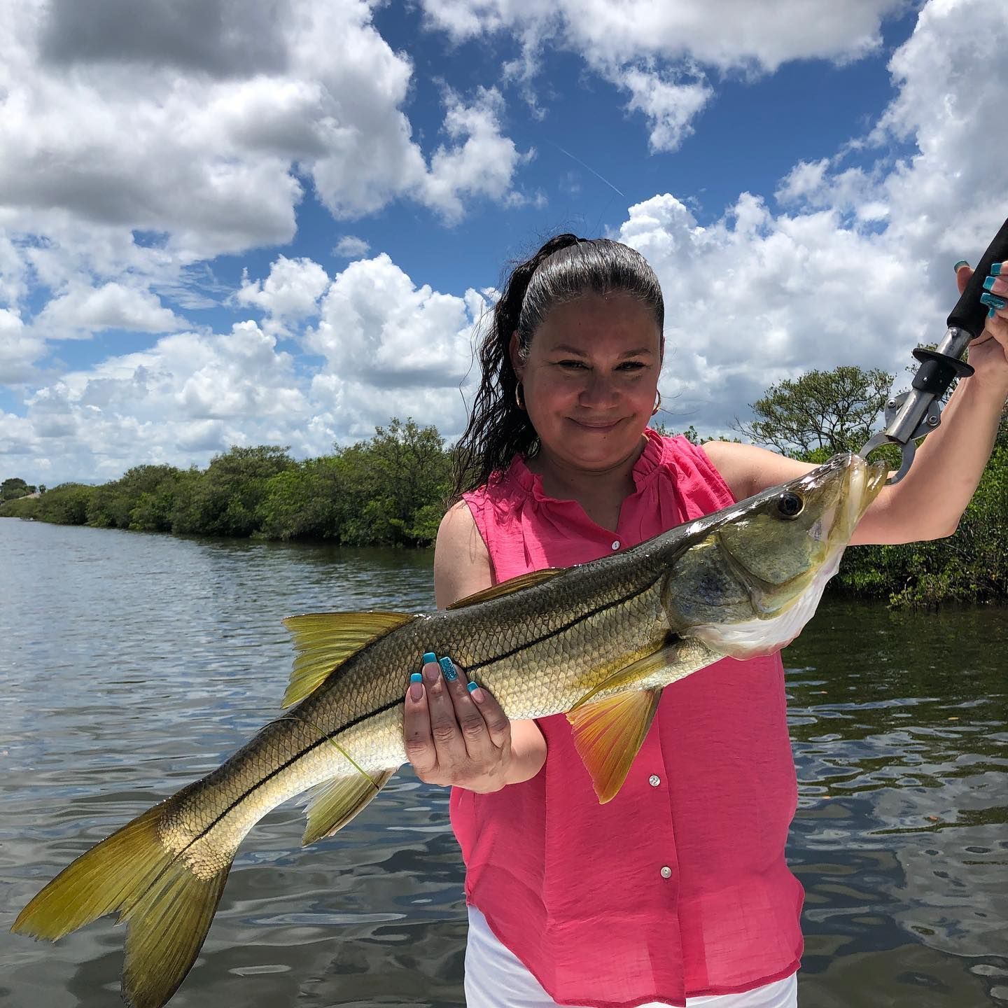 Woman on boat holds up a large fish, smiling, in a canal under a cloudy sky.