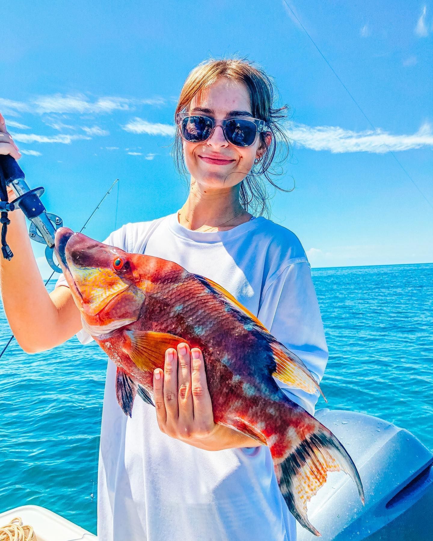 Woman on a boat holding a colorful fish, smiling at the camera, blue ocean background.