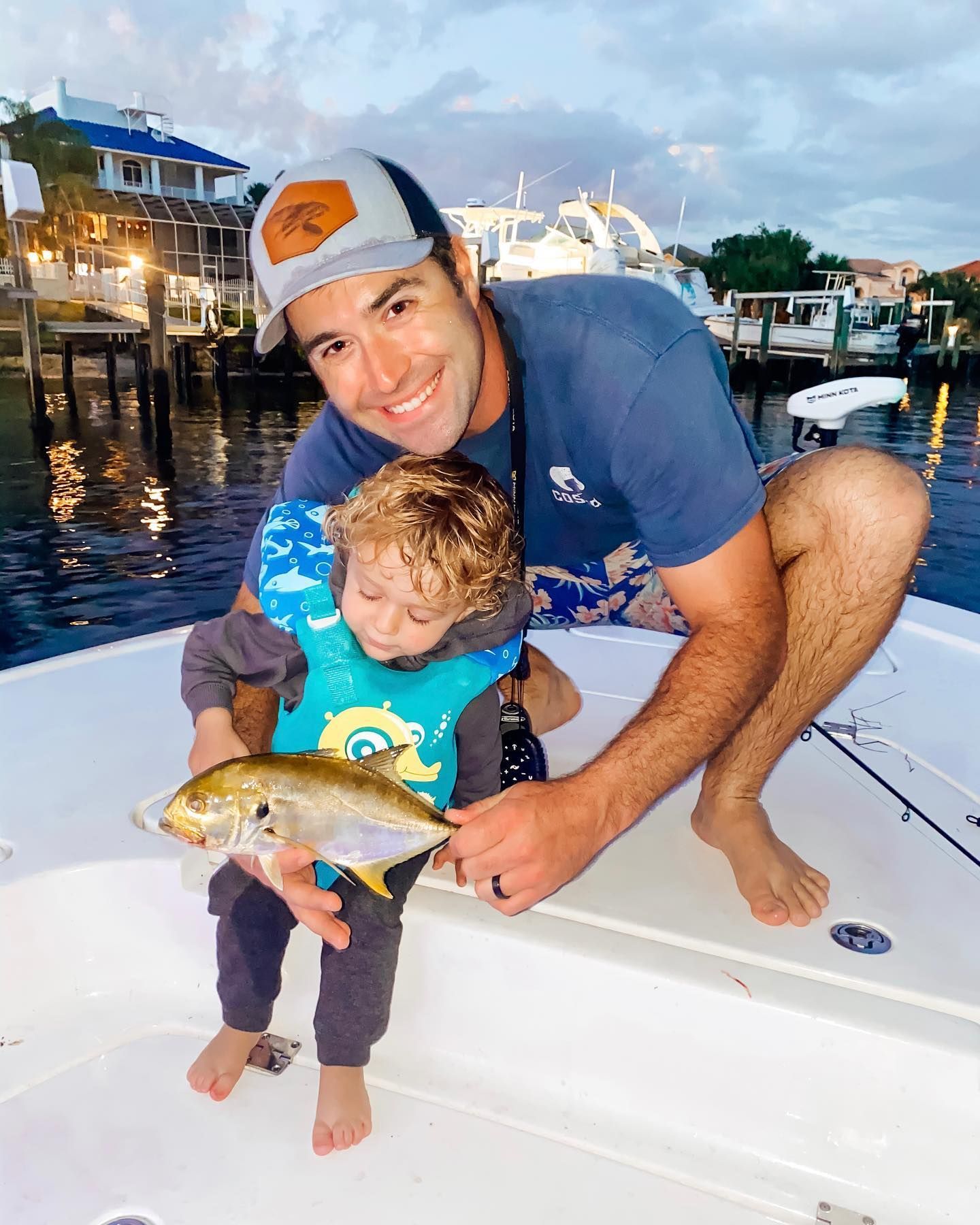 Man and child smile, holding a fish on a boat. Waterside setting.