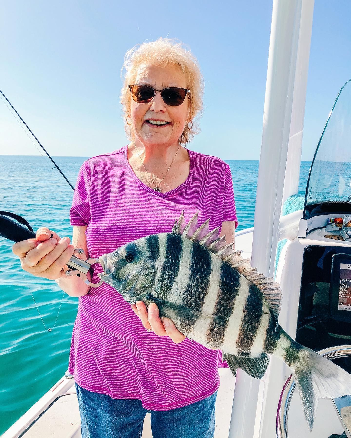 Woman on boat holds up a striped fish she caught. Smiling in sunglasses, blue water.
