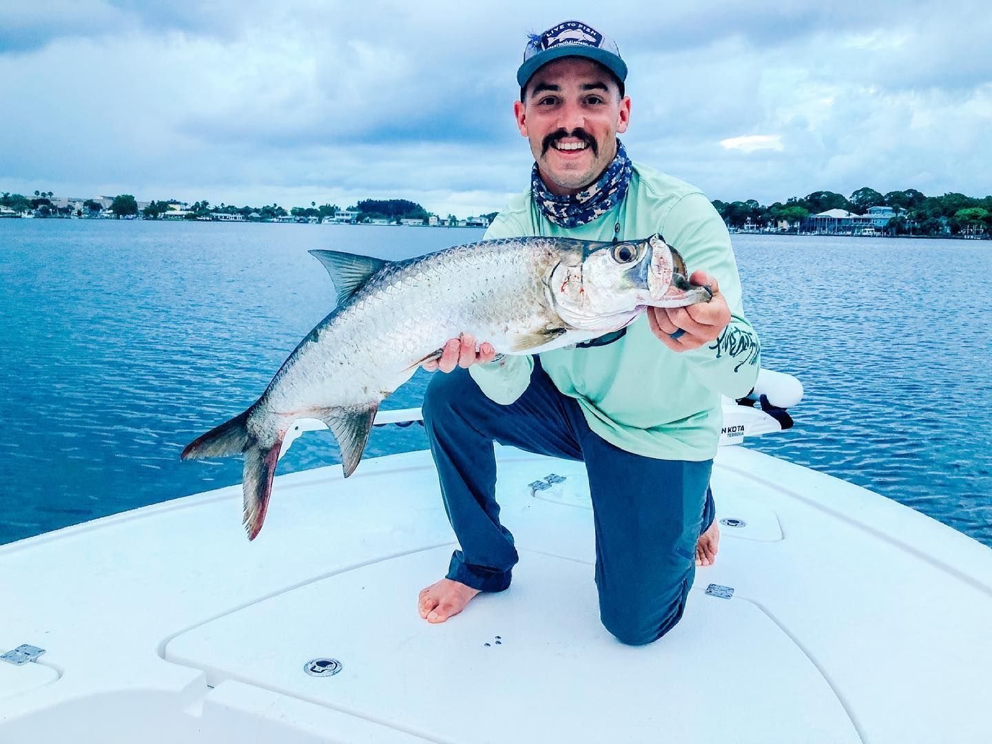 Man kneels on a boat, holding a silver tarpon. Overcast day, water behind him.