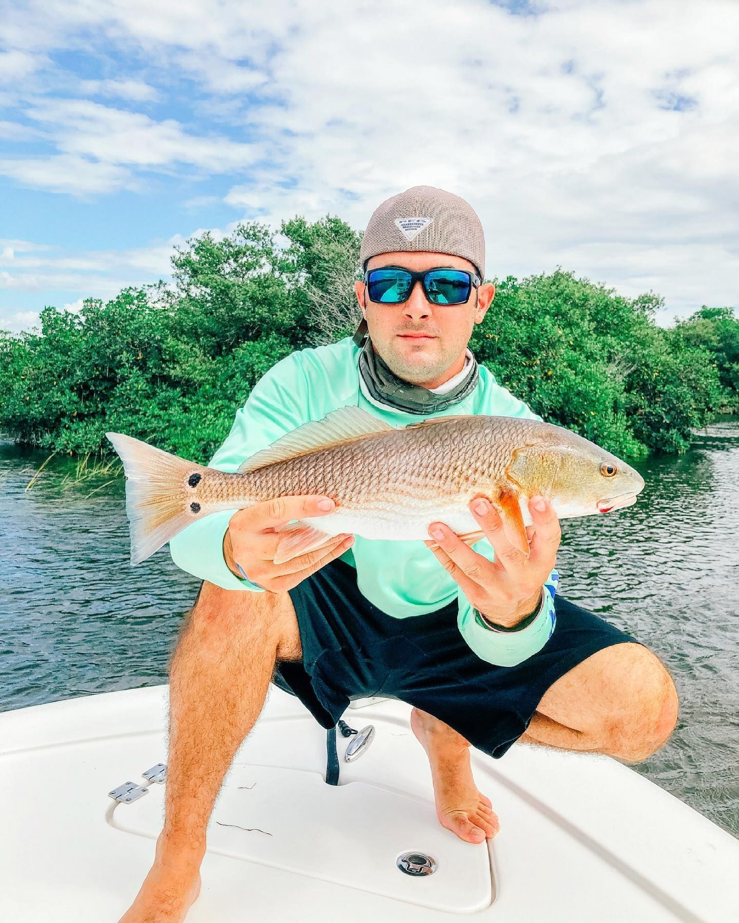 Man kneeling on boat holds up redfish, near mangroves, wearing sunglasses and a hat.