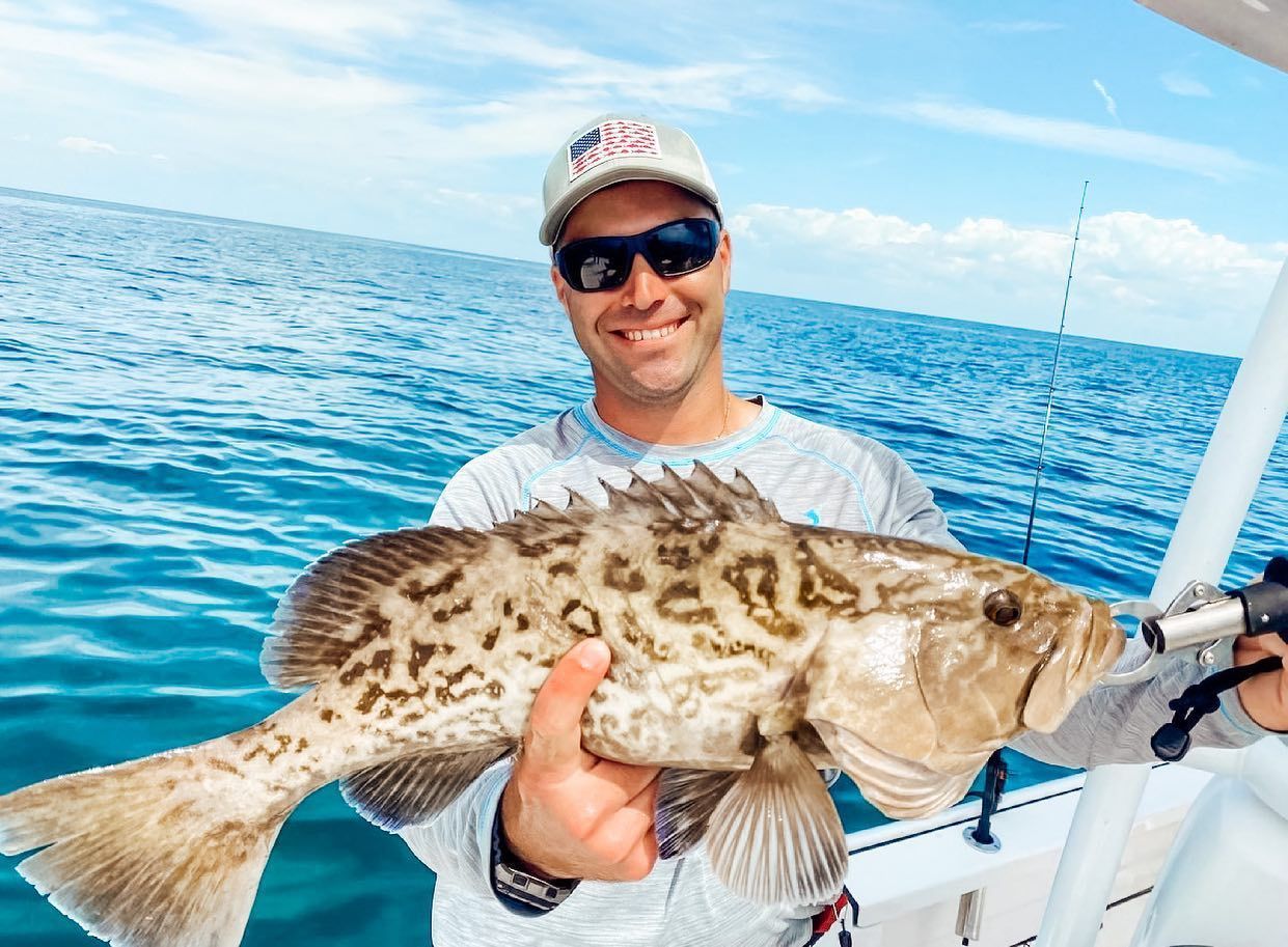 Man smiles, holding a large, spotted fish on a boat in the ocean.