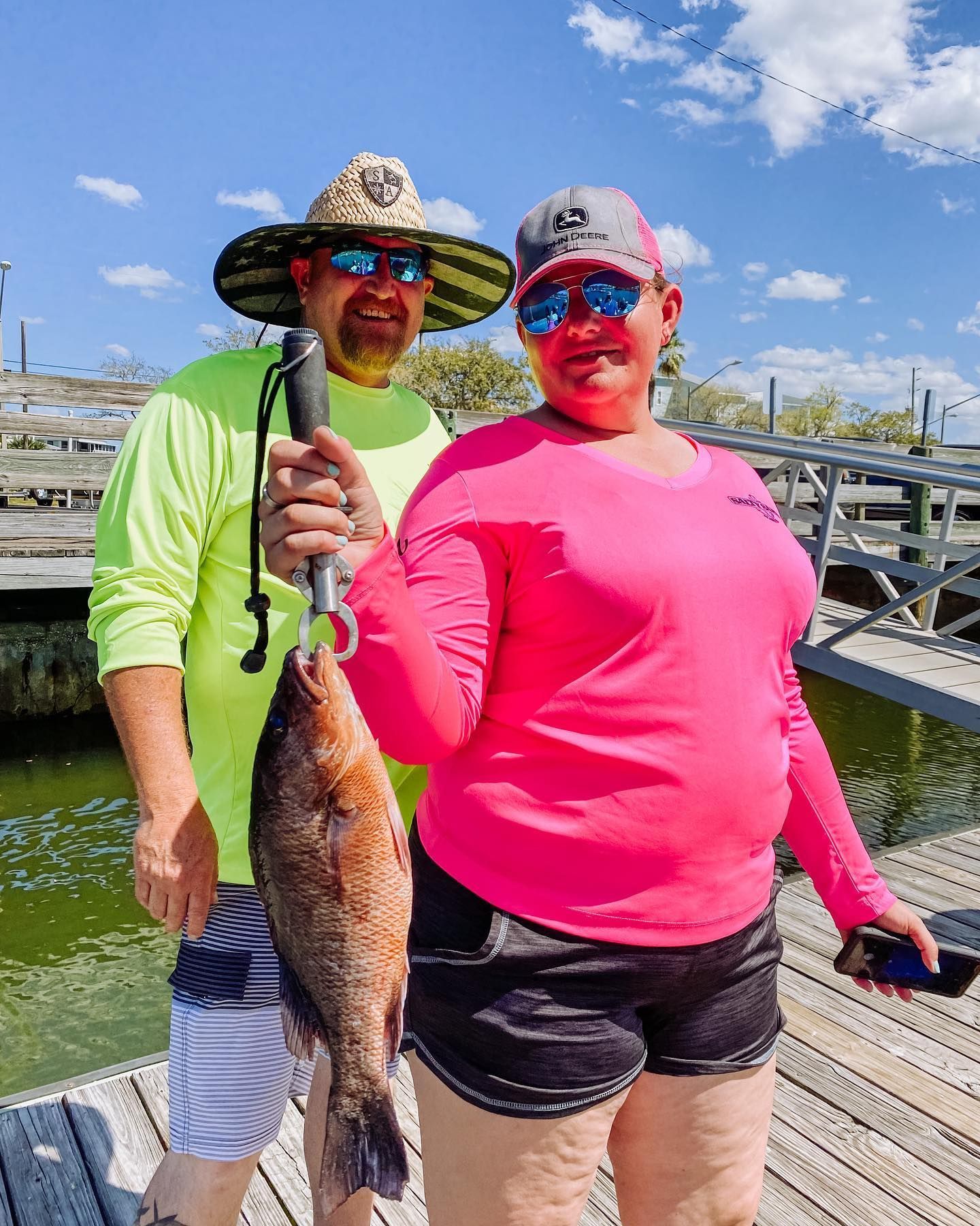 A couple smiles, holding a fish at a dock. Man in neon shirt, woman in pink. Sunny day.