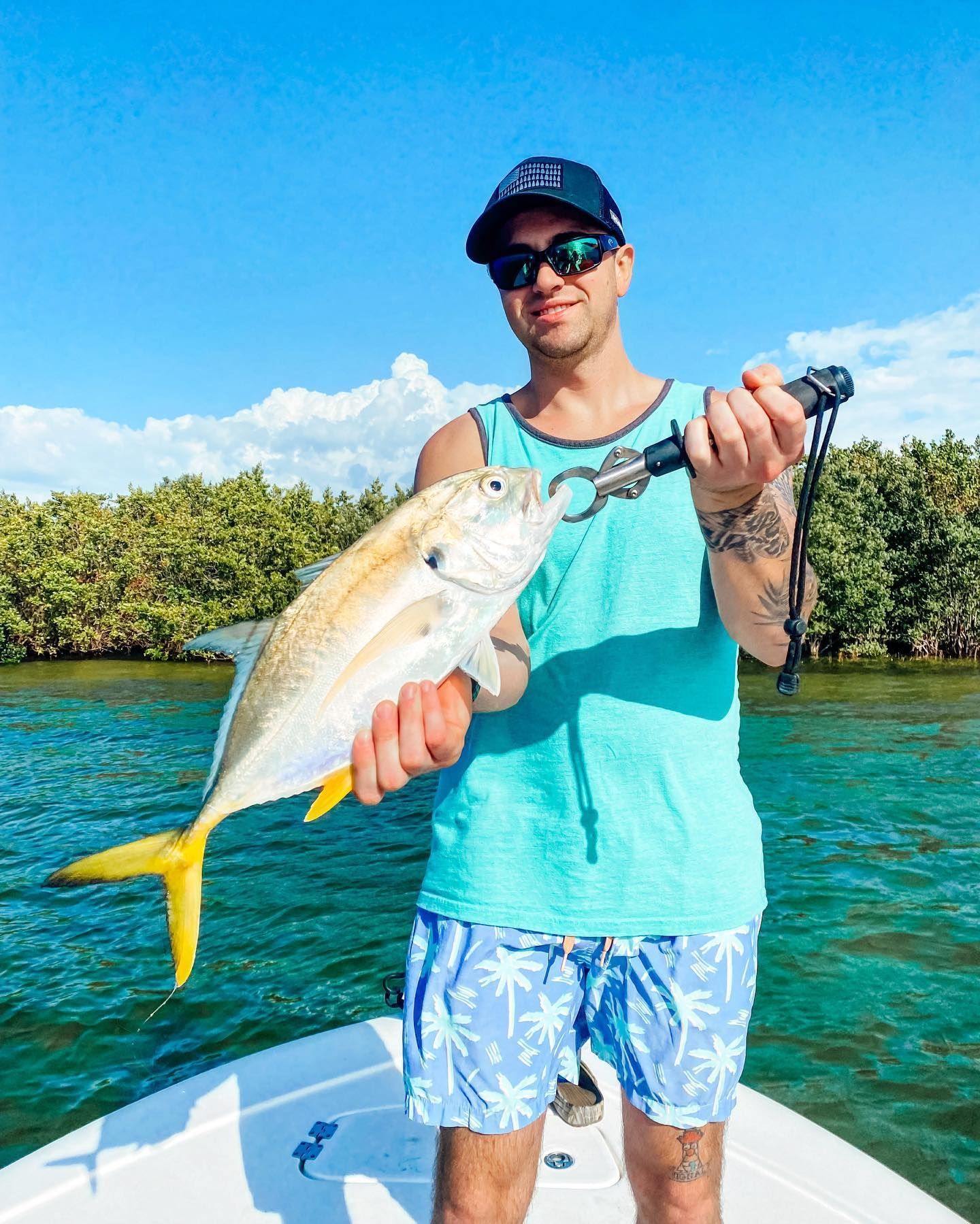 Man on boat holding a yellow jack fish. Blue sky, green water, mangroves.