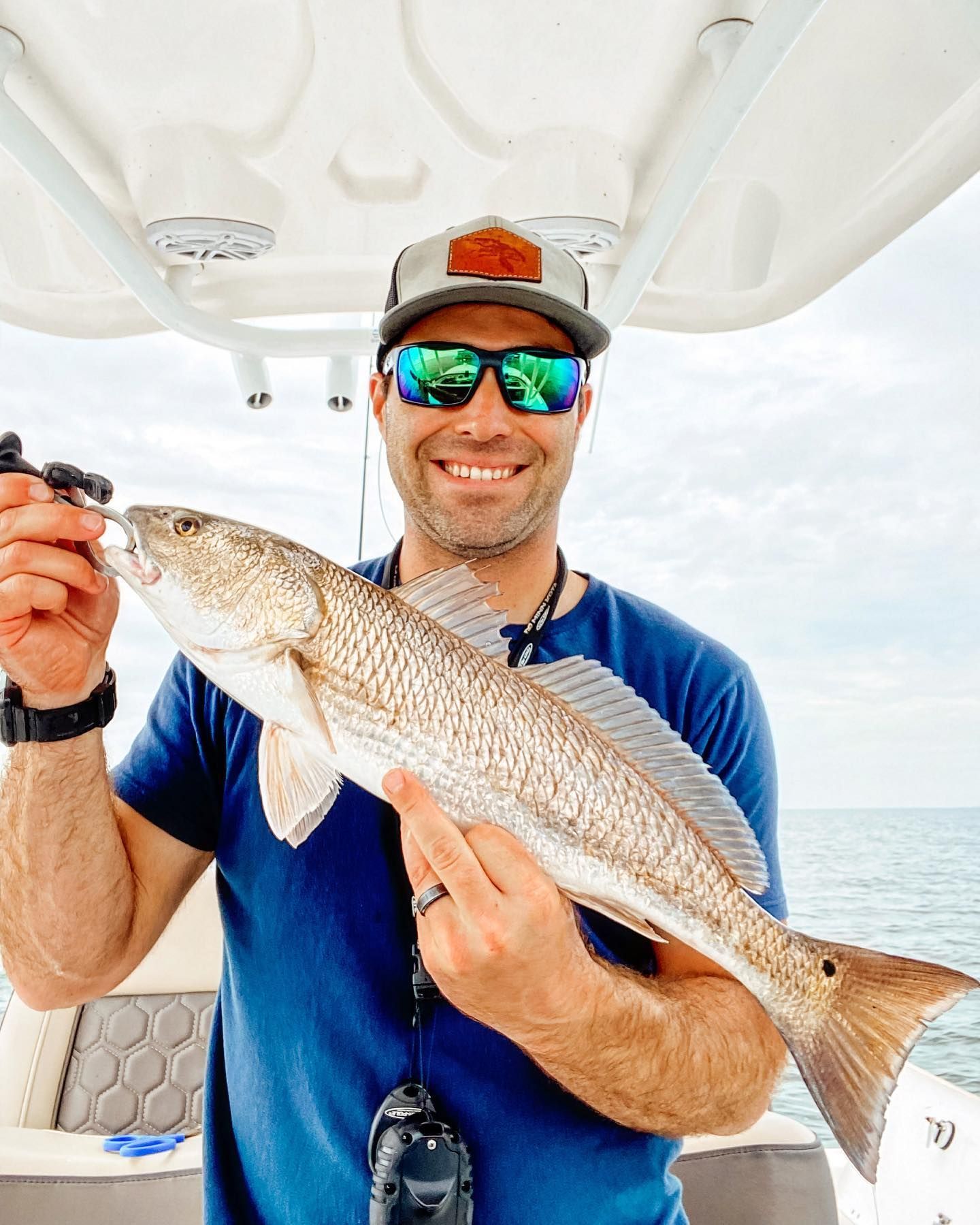 Man on a boat holding a redfish, smiling. Wearing sunglasses, a hat, and a blue shirt. Ocean background.