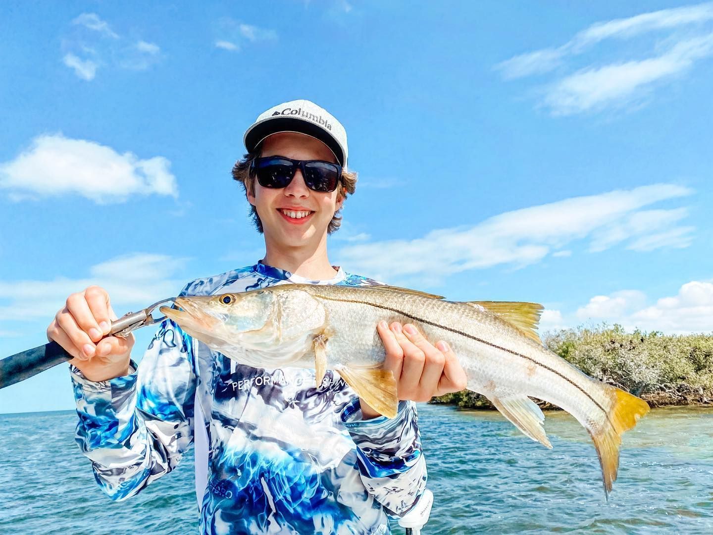 Young man smiling, holding a fish on a boat with blue sky and water.