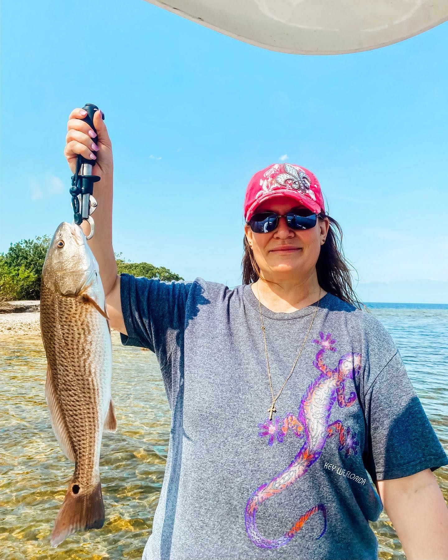 Woman in red hat and sunglasses holds up a reddish fish on a sunny day near the water.