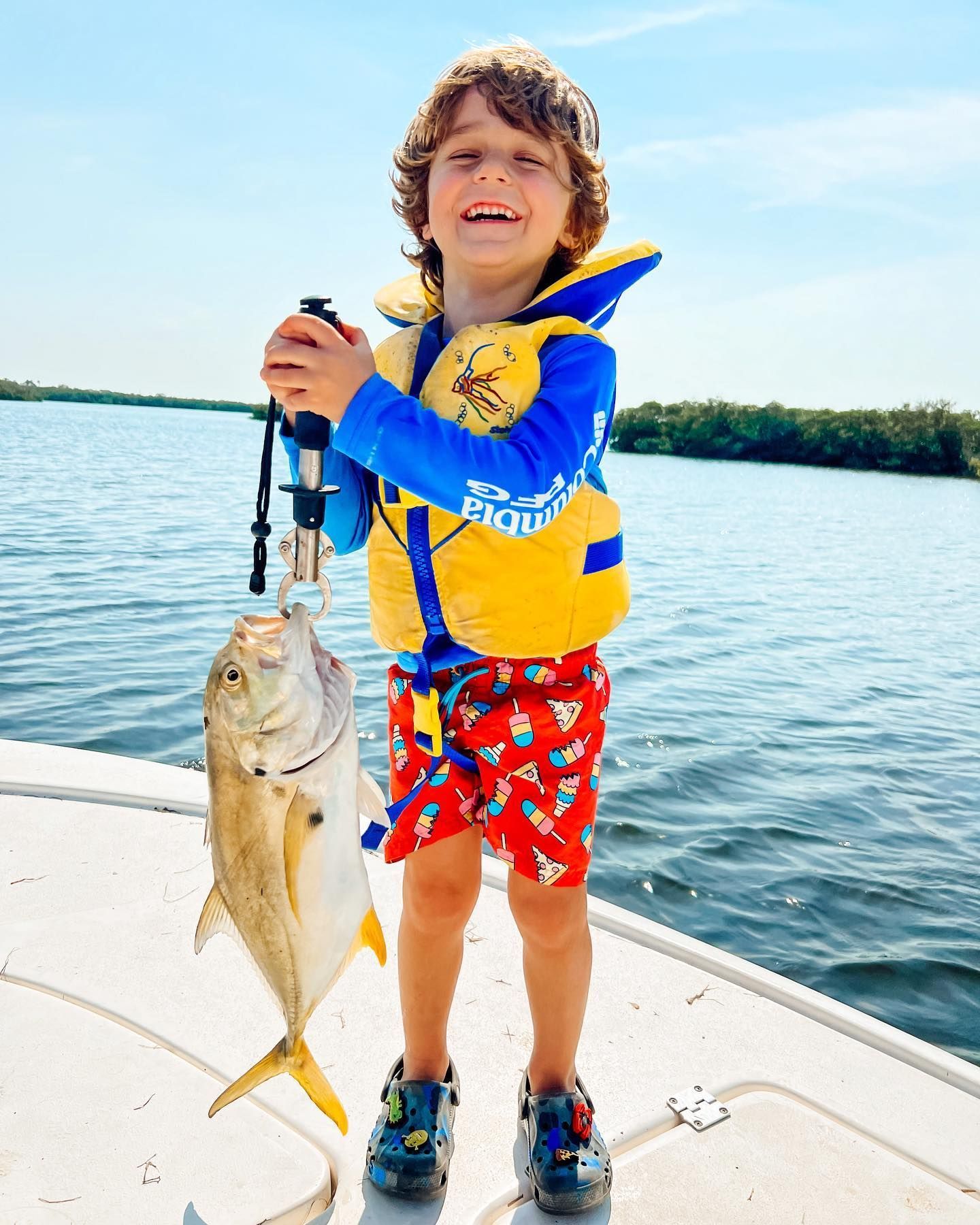 Boy on a boat holding up a fish he caught; he's smiling, wearing a life vest, blue shirt, and red shorts.