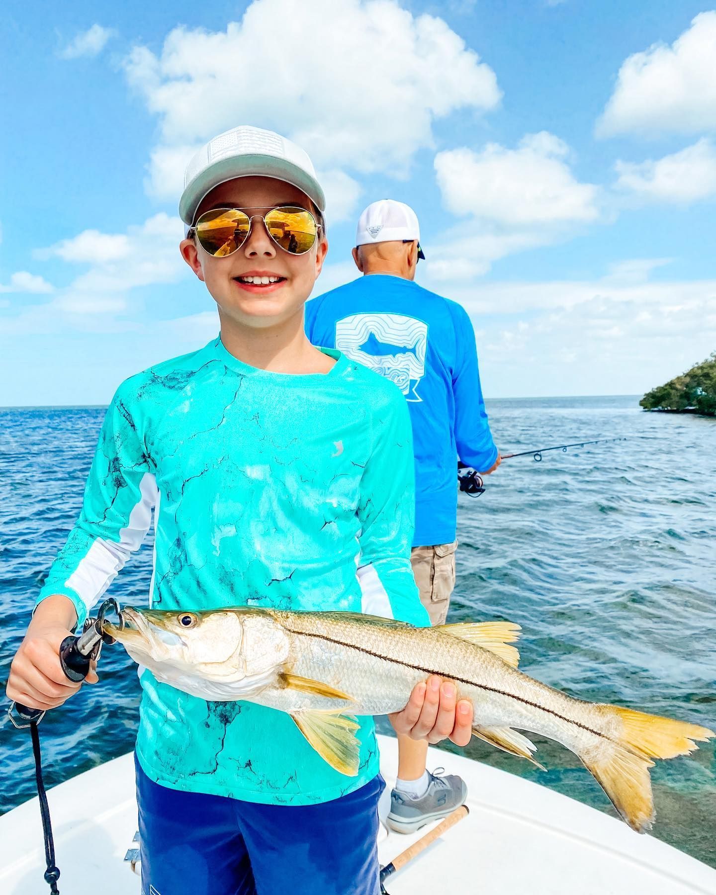 Boy smiling, holding a fish on a boat, blue water, man in background.