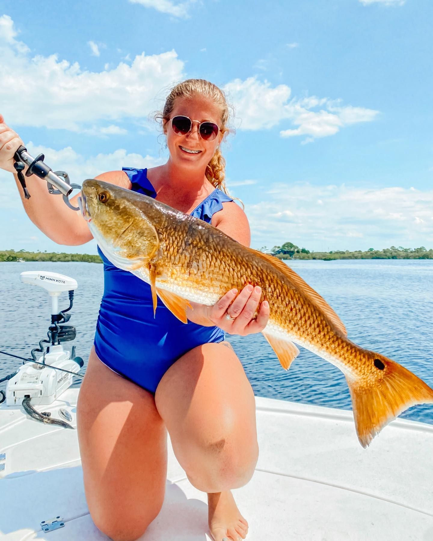 Woman kneeling on boat holds up a large redfish, blue water and sky.