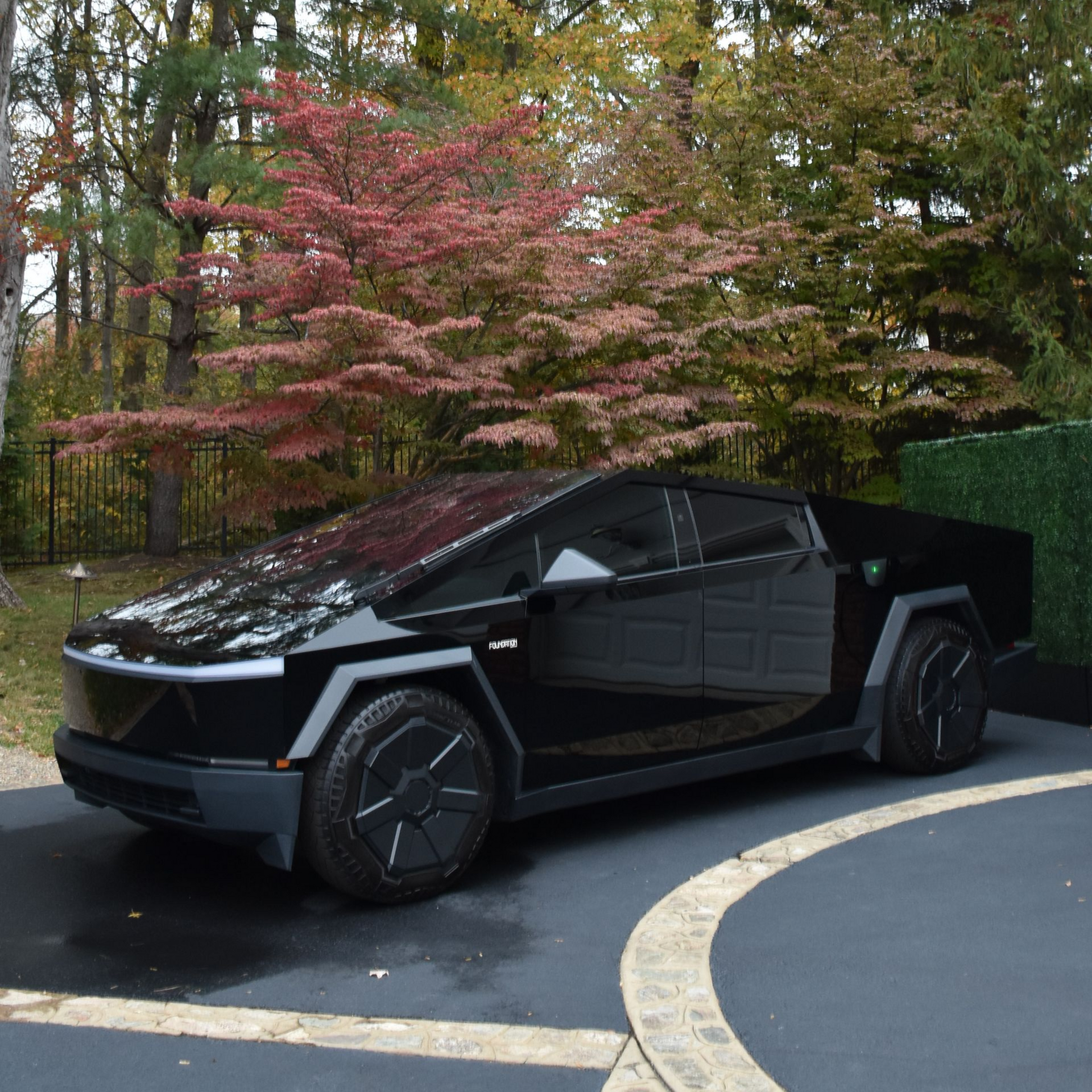 A person washes a silver car with soapy foam. Black trailer and basketball hoop in the background.