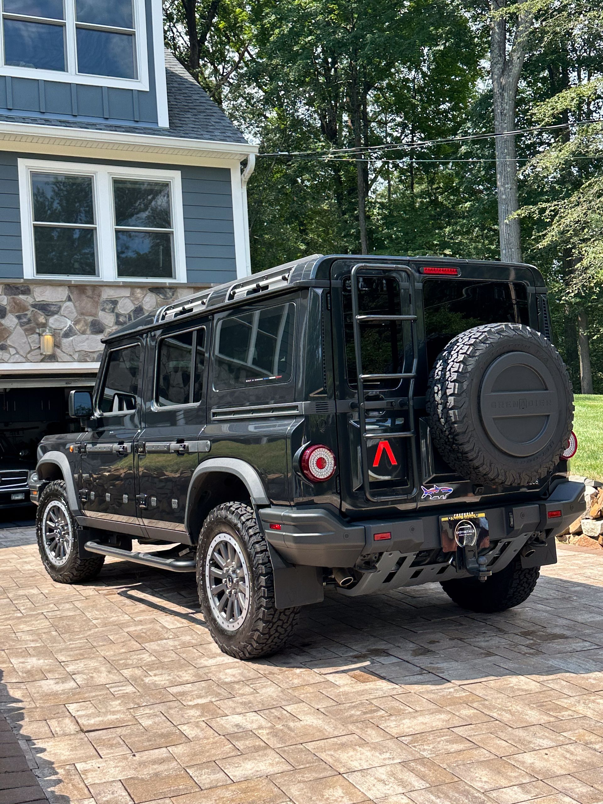 Black SUV parked on a brick driveway, next to a blue house. Features a rear ladder and spare tire.