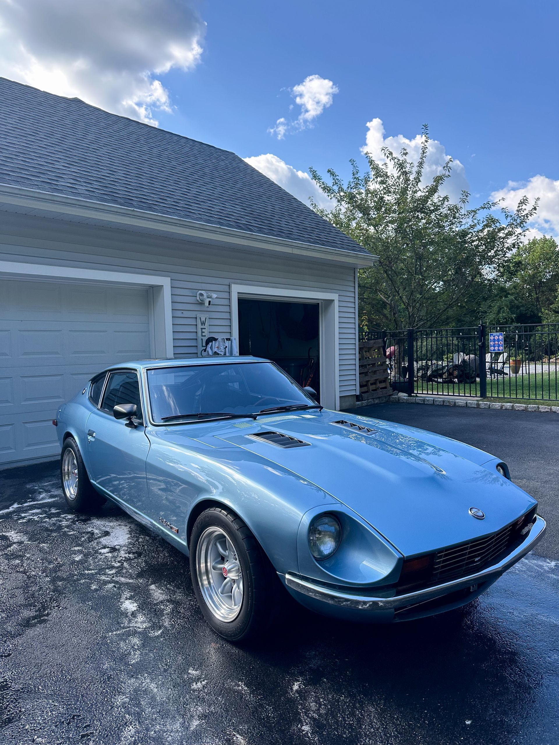 Light blue classic Datsun sports car parked near a garage on a sunny day.