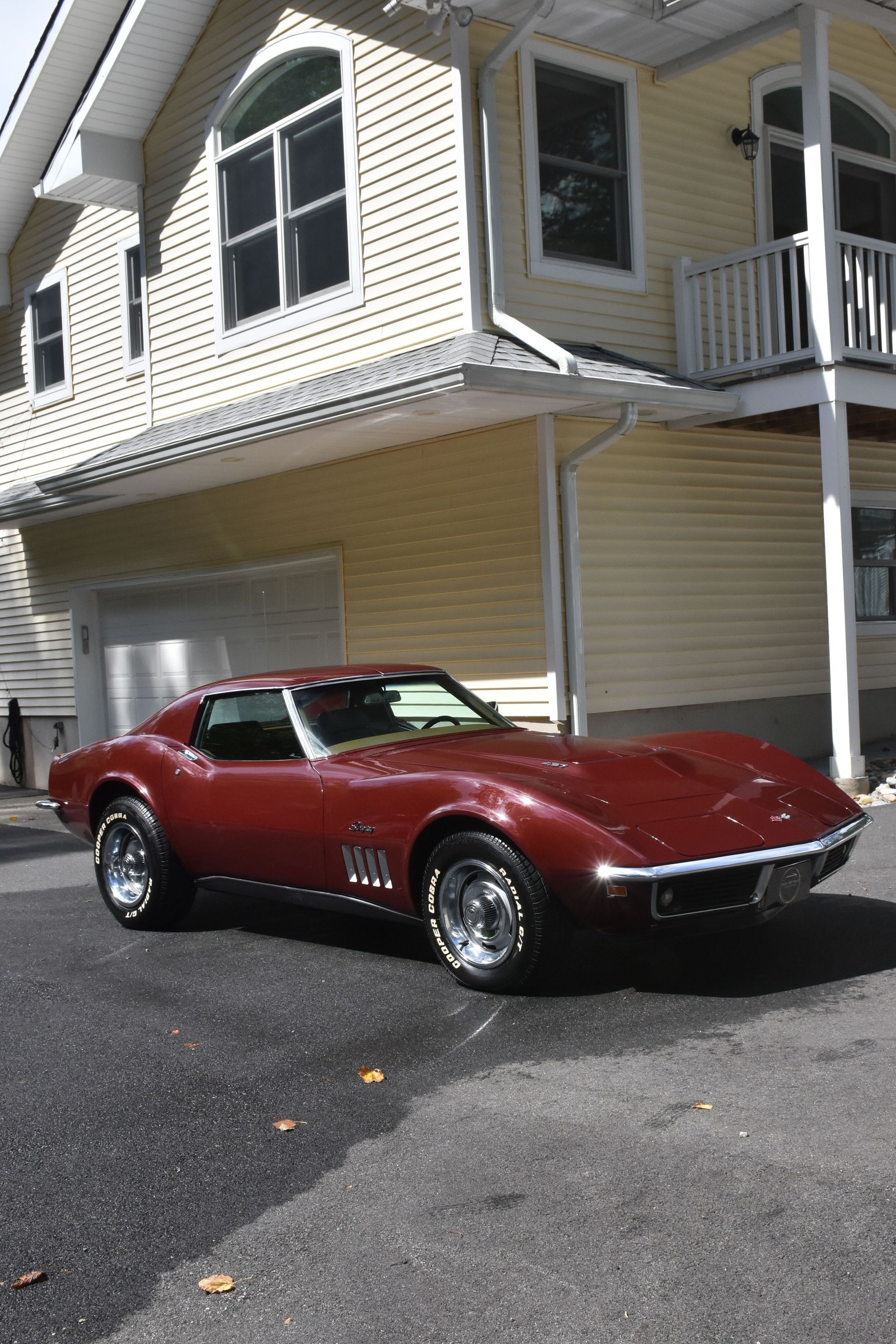 Maroon 1968 Chevrolet Corvette parked in front of a yellow house with a white garage.