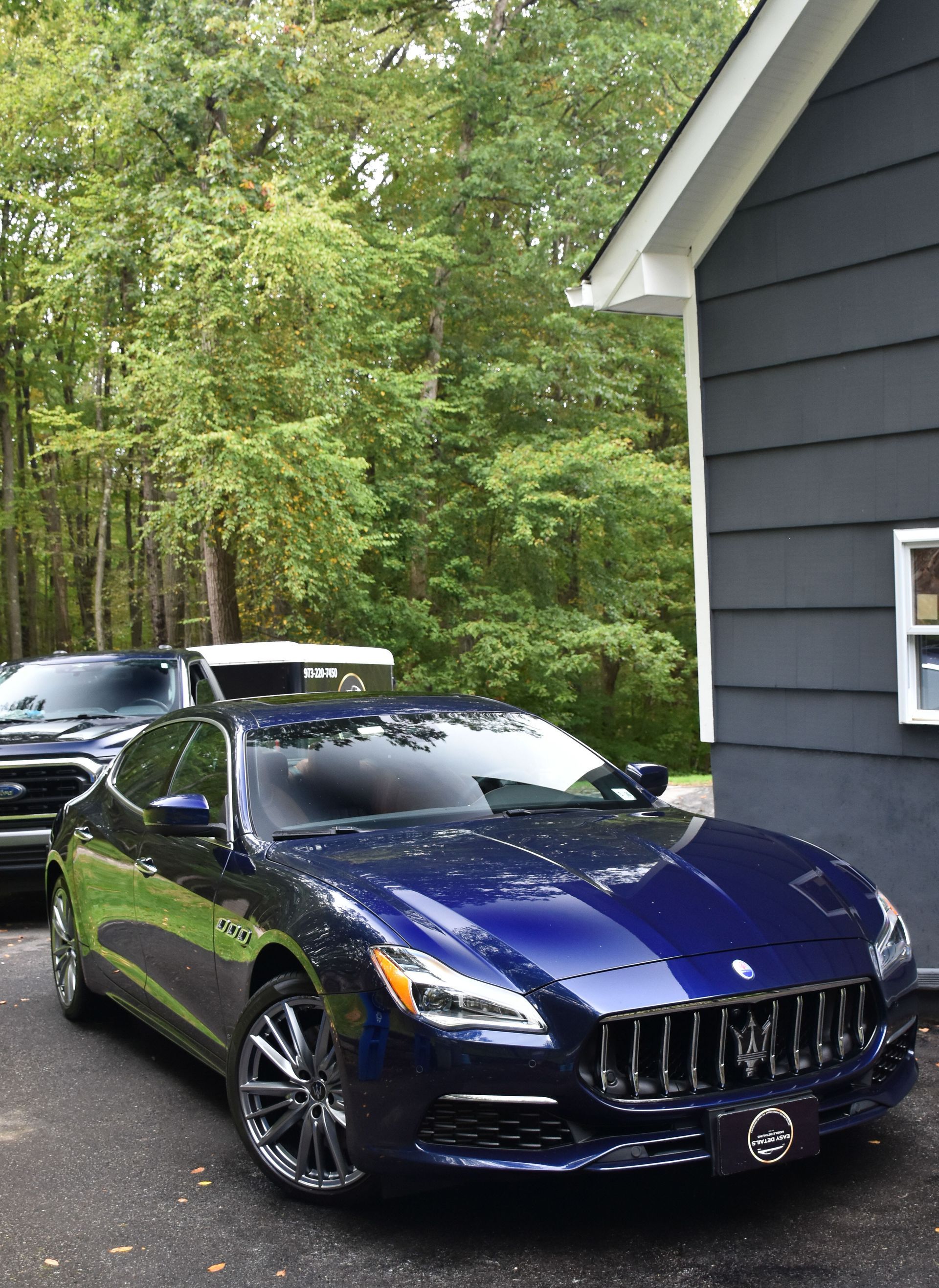 Blue Maserati sedan parked next to a gray house, with a dark-colored SUV visible behind it.