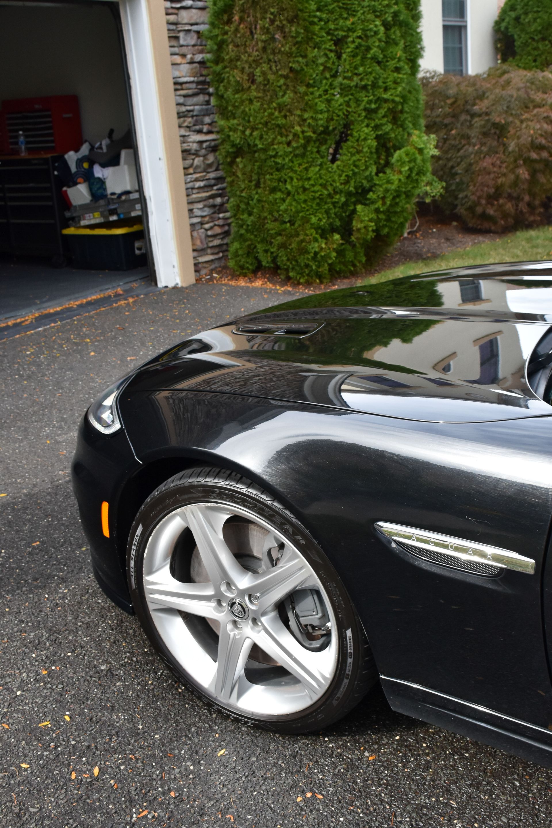 Black Jaguar car parked on asphalt driveway, in front of garage and shrubs. Shiny paint.