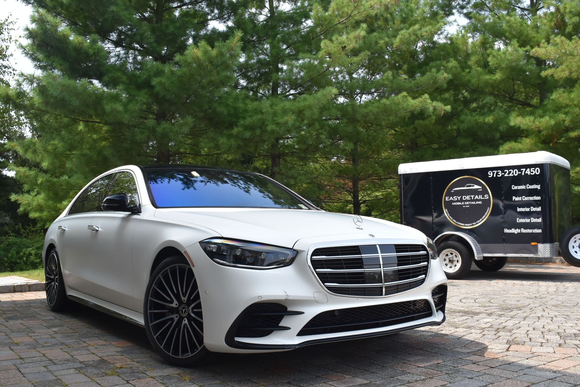 White Mercedes sedan parked next to a black and white trailer. Green trees in the background.