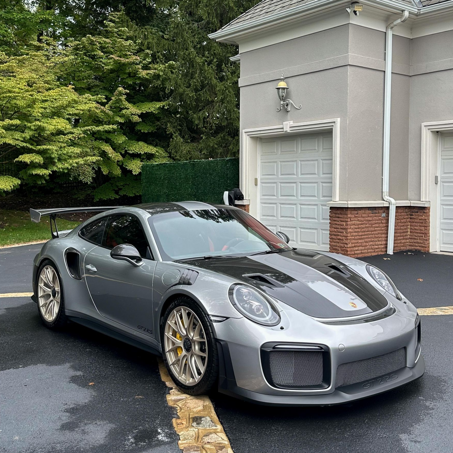 Silver Porsche 911 parked in front of a house, dark stripe on hood, gold wheels, wet pavement.
