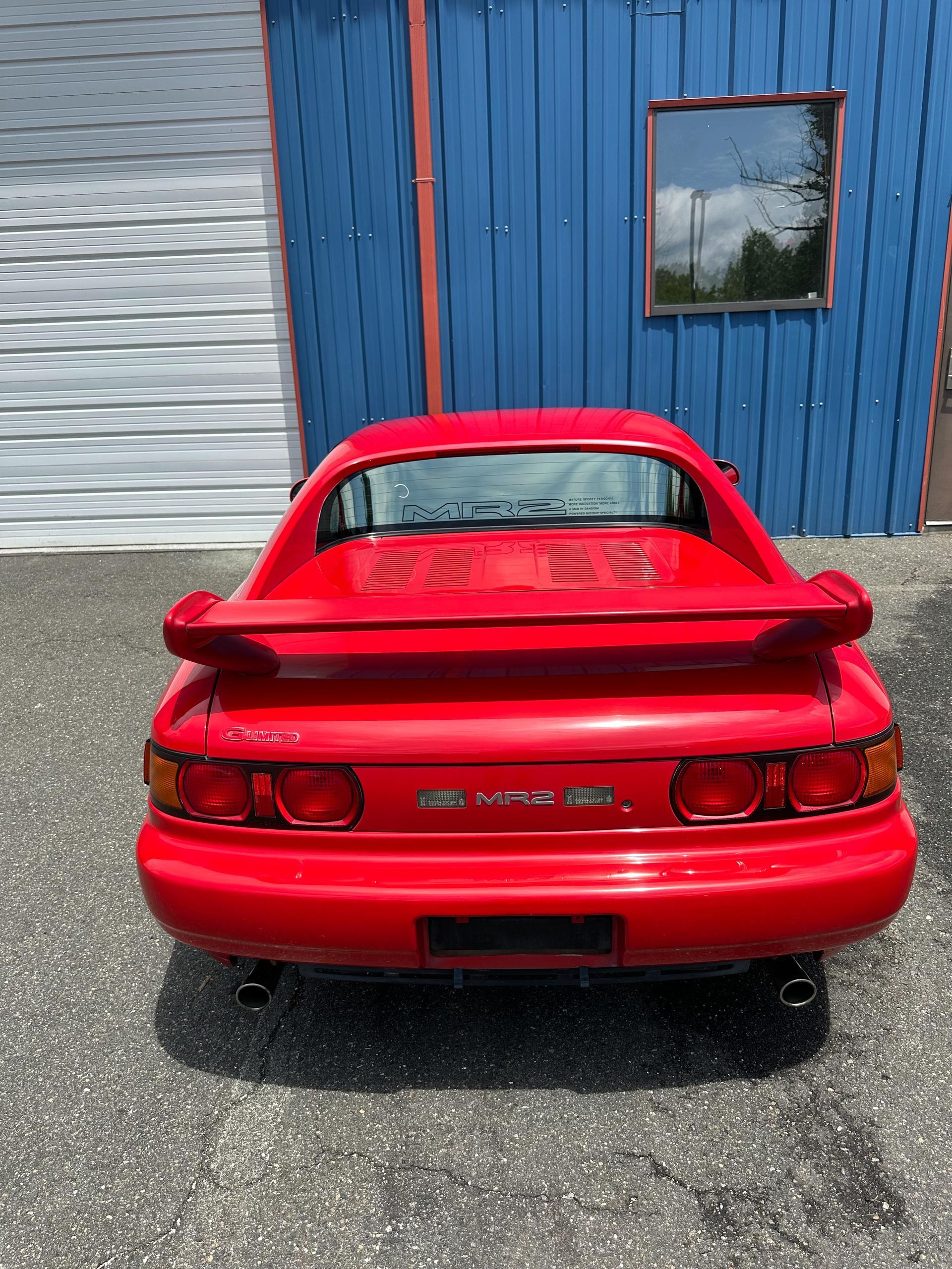 Red Toyota MR2 sports car parked in front of a blue building with a small window.