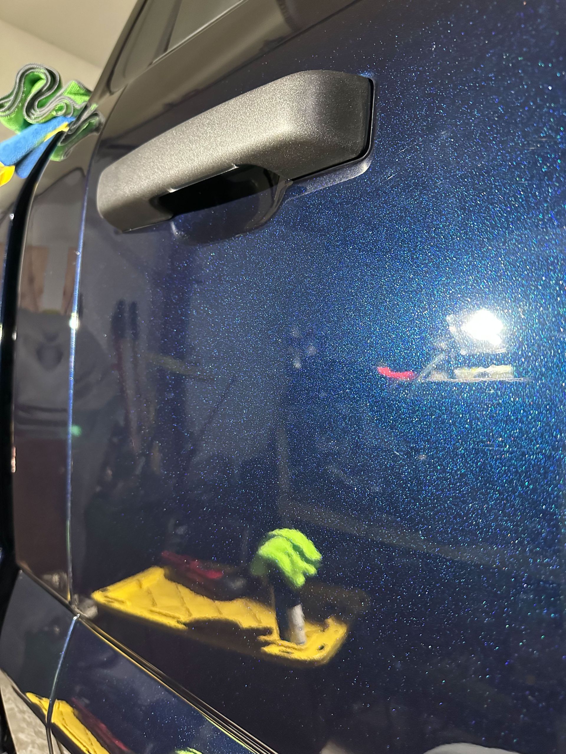 Blue car door with glitter-like paint; black handle; reflection of a person in the paint holding a green glove.