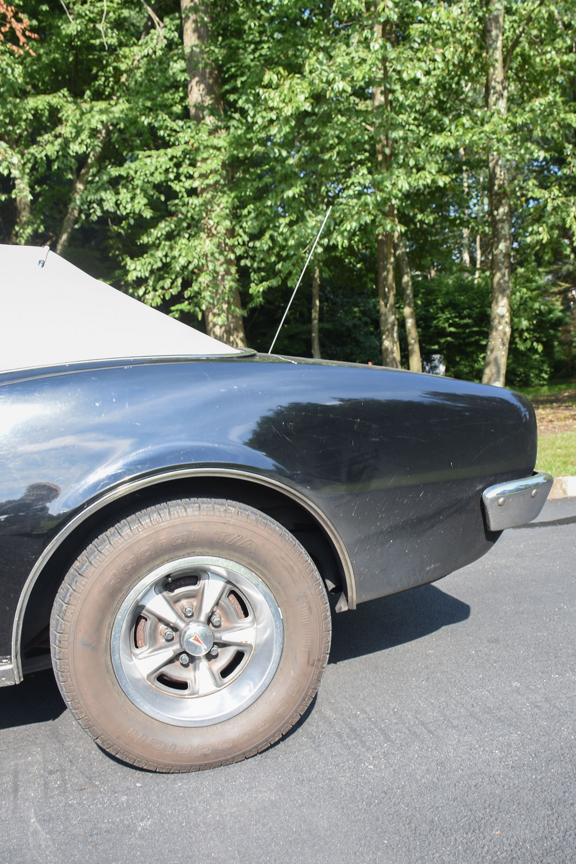 Black classic car's rear quarter panel and wheel. Trees in background, car is parked on asphalt.