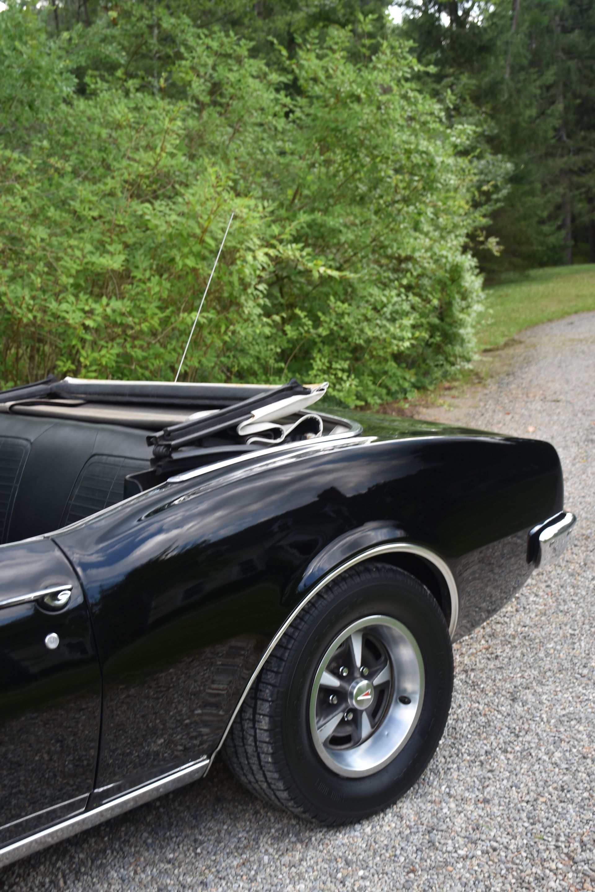 Black classic convertible car parked on a gravel road, trees in the background.