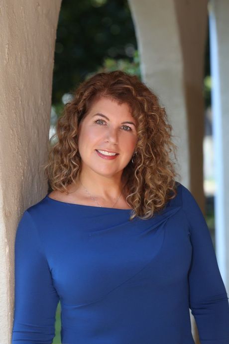 A woman in a blue shirt is leaning against a wall and smiling.