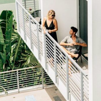 Woman and man on a balcony with white railings, looking at each other. Lush green plants in the background.