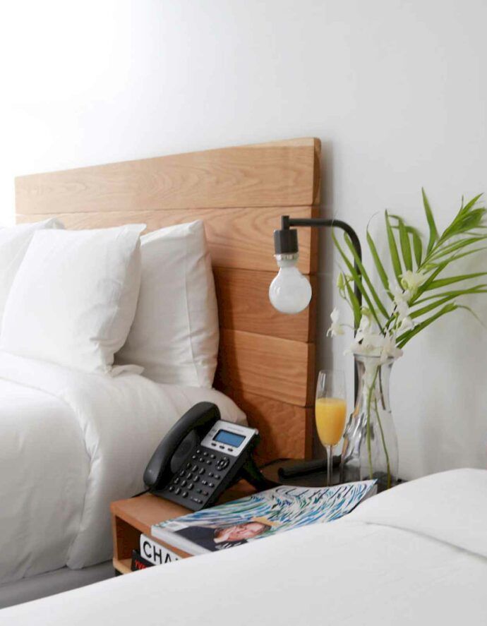 Bedroom scene: bed with white linens, wooden headboard, bedside table with lamp, phone, magazine, and flowers.