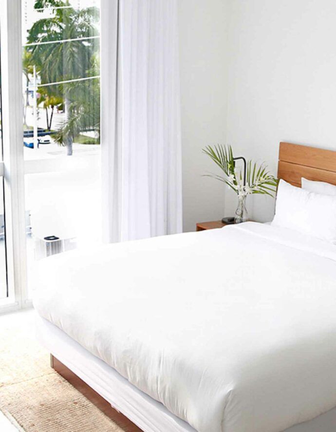 White bedroom with bed, wooden headboard, window with curtains, and vase of leaves.