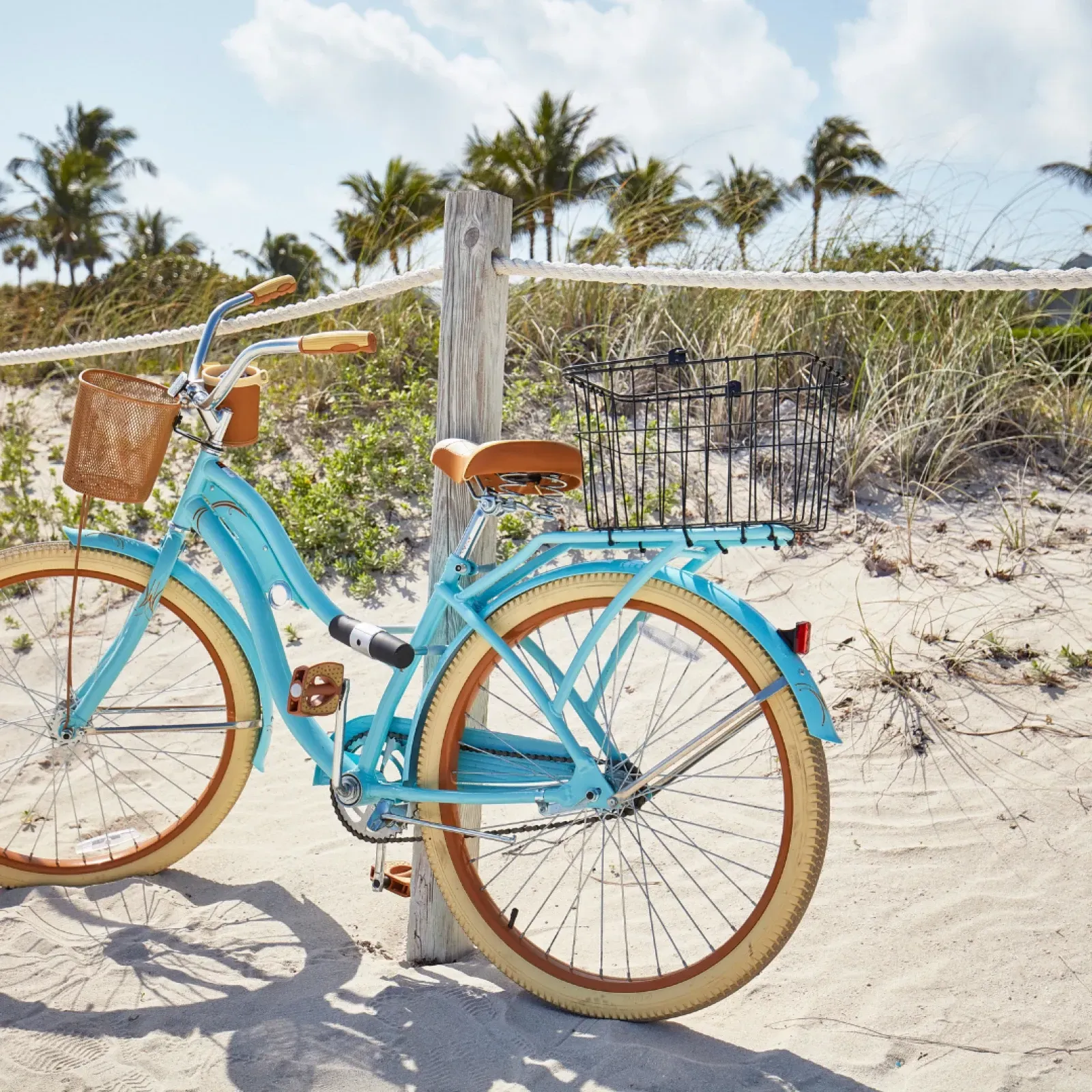 Light blue bicycle parked on a sandy beach, with a wooden fence and palm trees in the background.